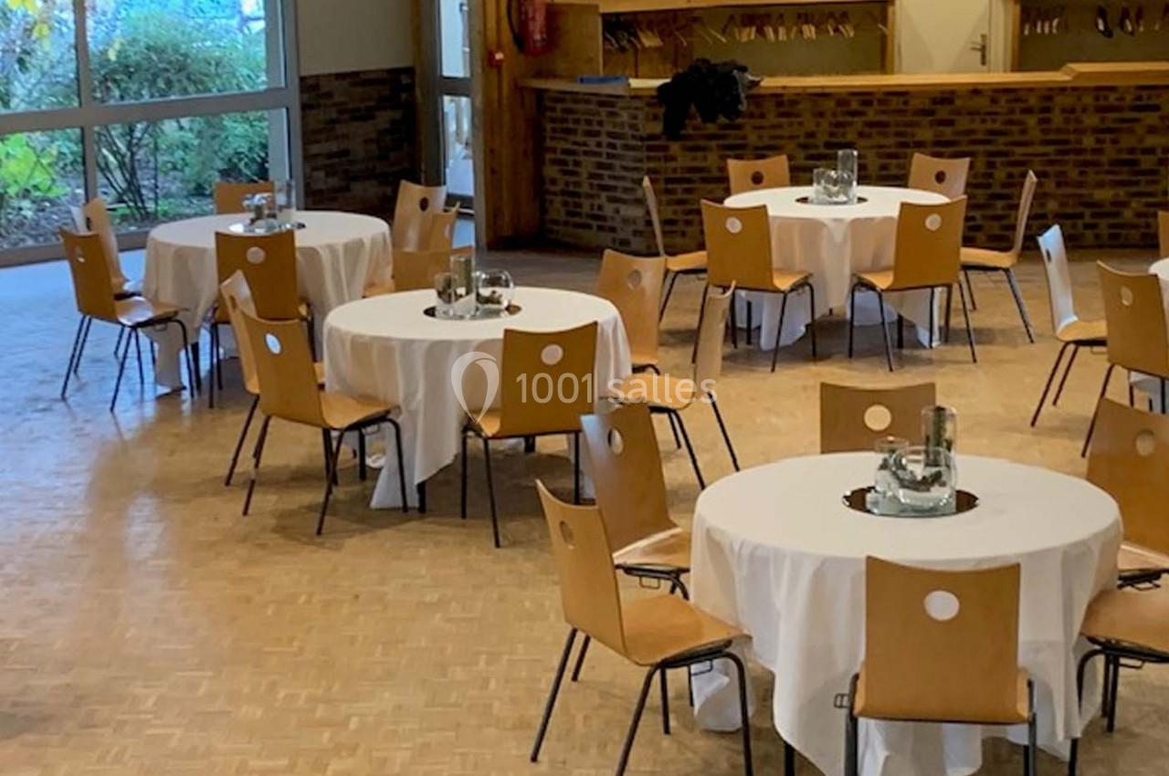 Salle avec des tables rondes dressées de nappes blanches, entourées de chaises en bois, près de grandes fenêtres.