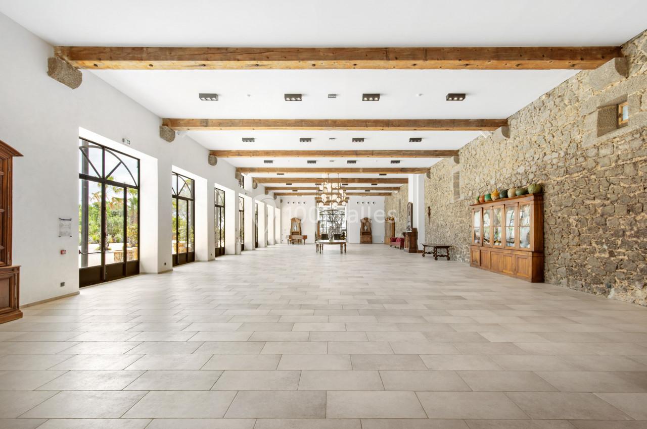 Grande salle lumineuse avec murs en pierre, poutres apparentes, carrelage clair et grandes fenêtres donnant sur l'extérieur.