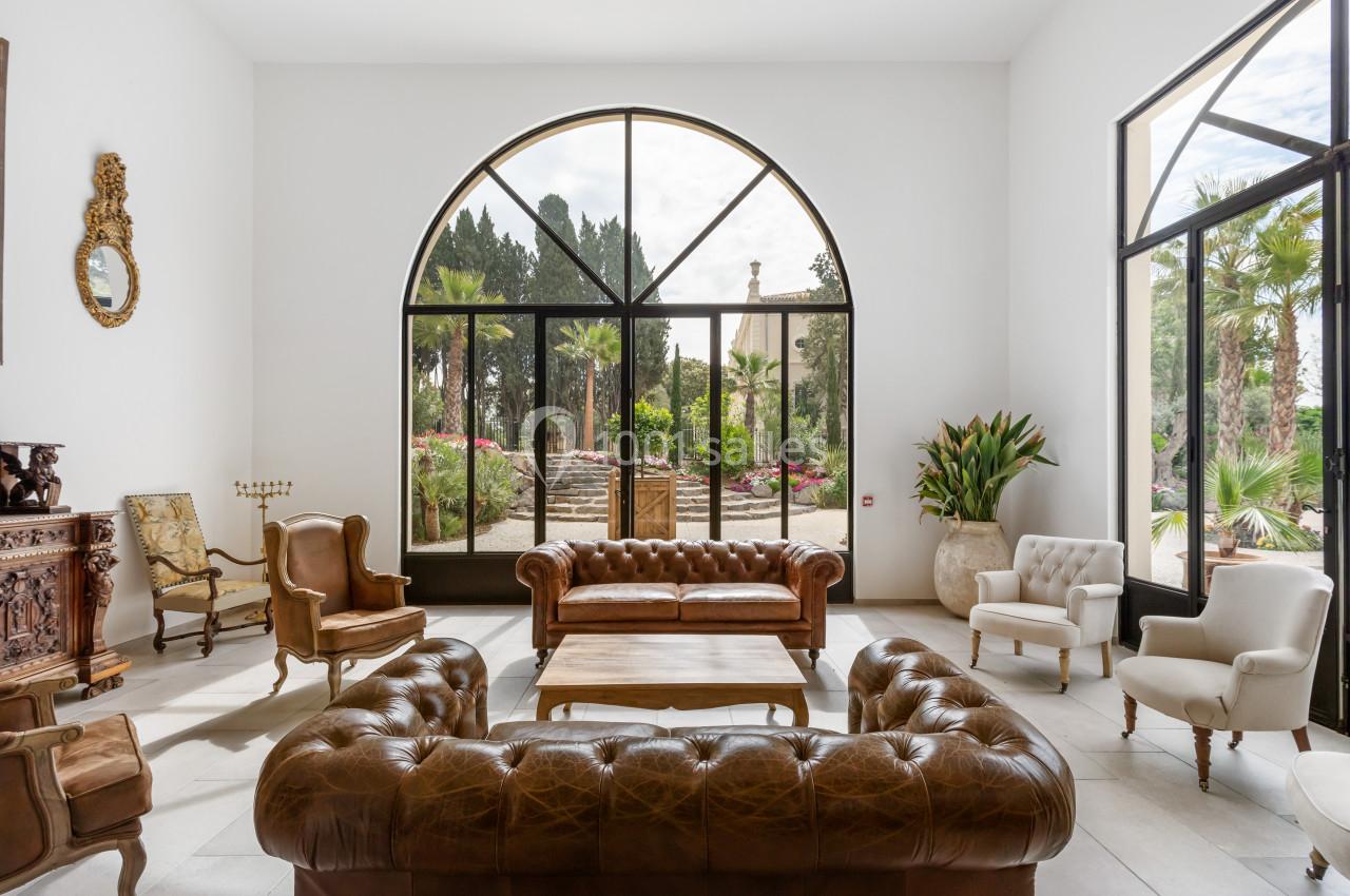 Salon lumineux avec canapés en cuir, fauteuils classiques et vue sur un jardin arboré à travers de grandes baies vitrées.