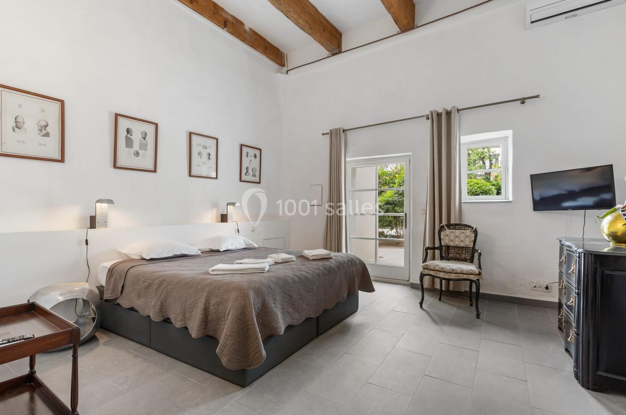 Chambre lumineuse avec lit double, murs blancs, poutres apparentes, fenêtres donnant sur un jardin et mobilier classique.