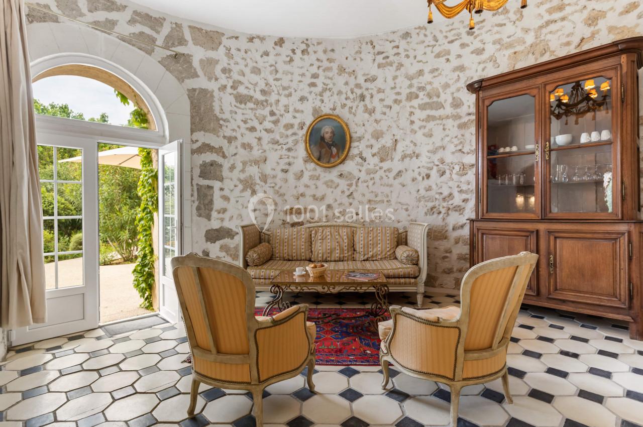 Salon lumineux avec murs en pierre, canapé et fauteuils classiques, armoire en bois et porte-fenêtre donnant sur un jardin.