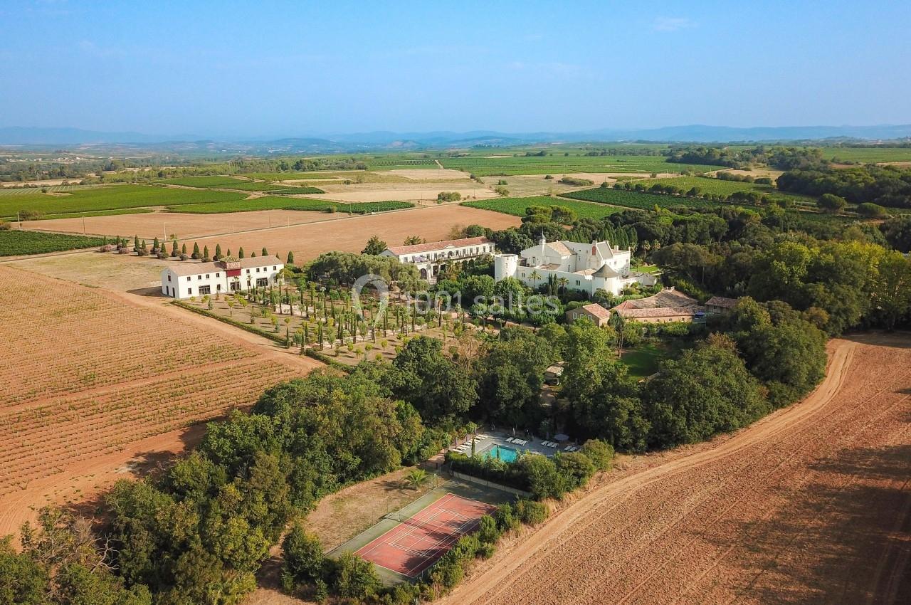 Vue aérienne d'un domaine entouré de vignes, avec jardins, piscine, courts de tennis et paysage rural en arrière-plan.