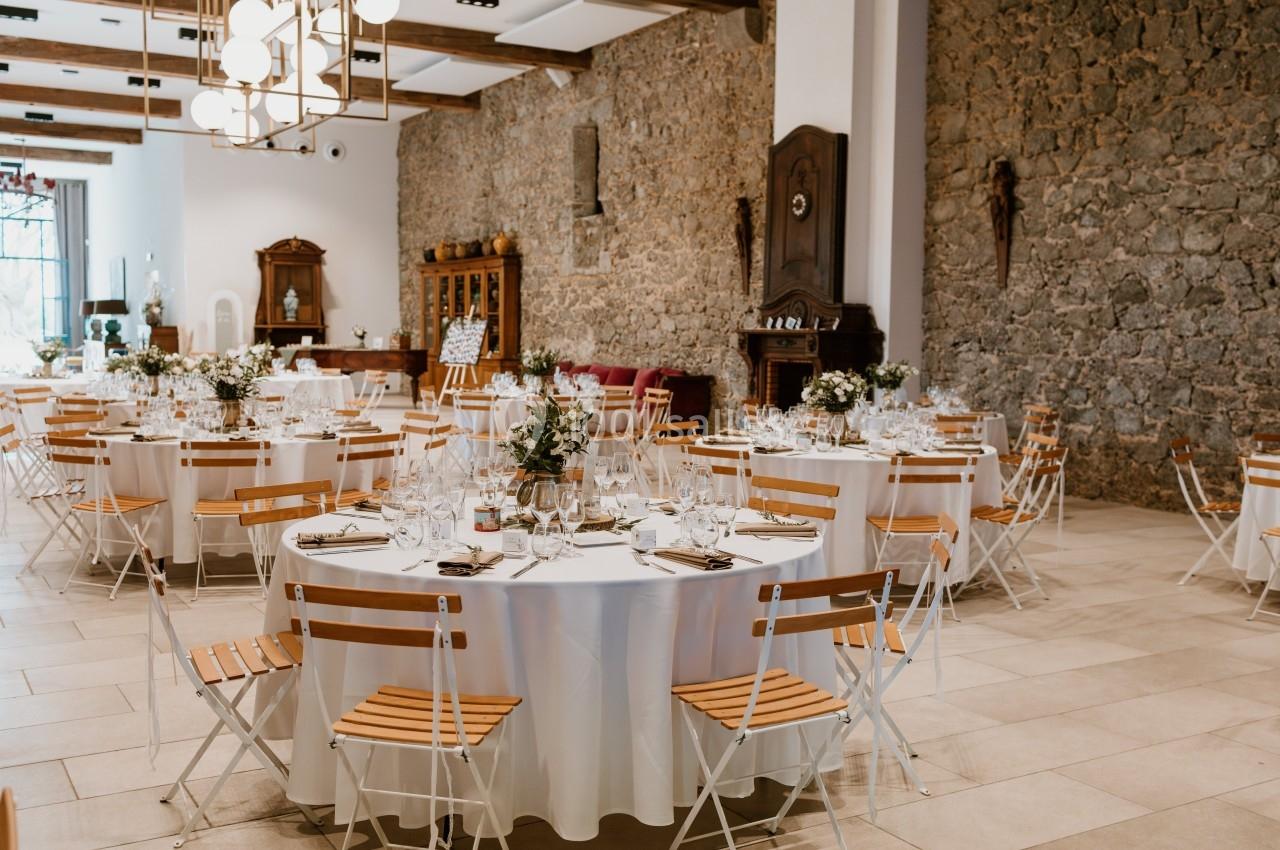 Salle de réception décorée avec des tables rondes dressées, chaises en bois et murs en pierre apparente.