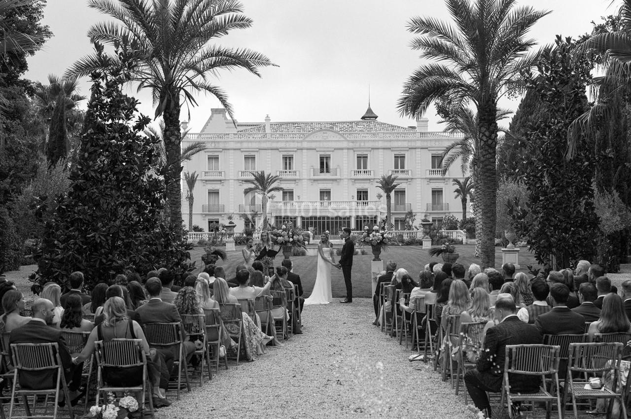 Cérémonie de mariage en plein air devant un manoir, avec des invités assis et des palmiers environnants.