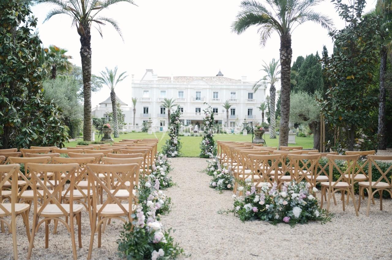 Allée centrale bordée de chaises en bois et de fleurs menant à un bâtiment blanc entouré de palmiers.