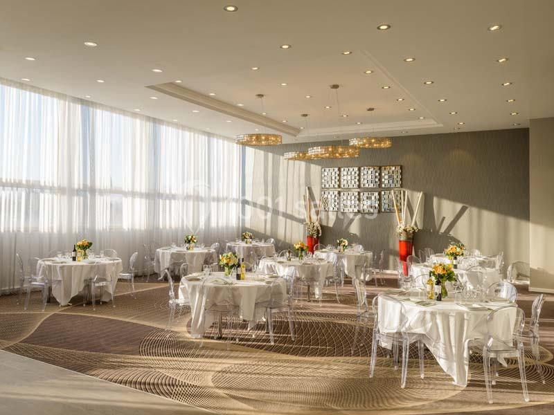 Salle lumineuse avec grandes fenêtres, tables rondes dressées avec nappes blanches, chaises transparentes et décor floral.