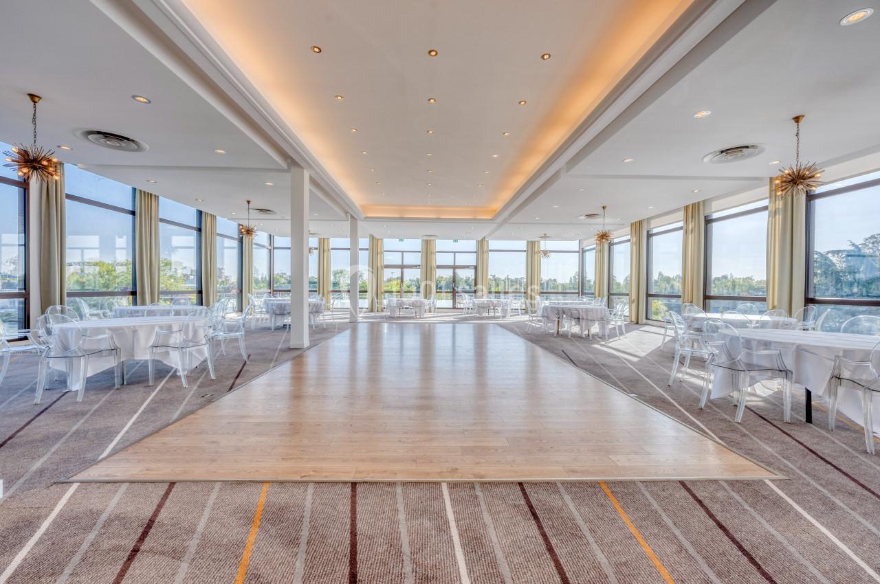 Salle lumineuse avec de grandes baies vitrées, tables rondes dressées et parquet central entouré de moquette.