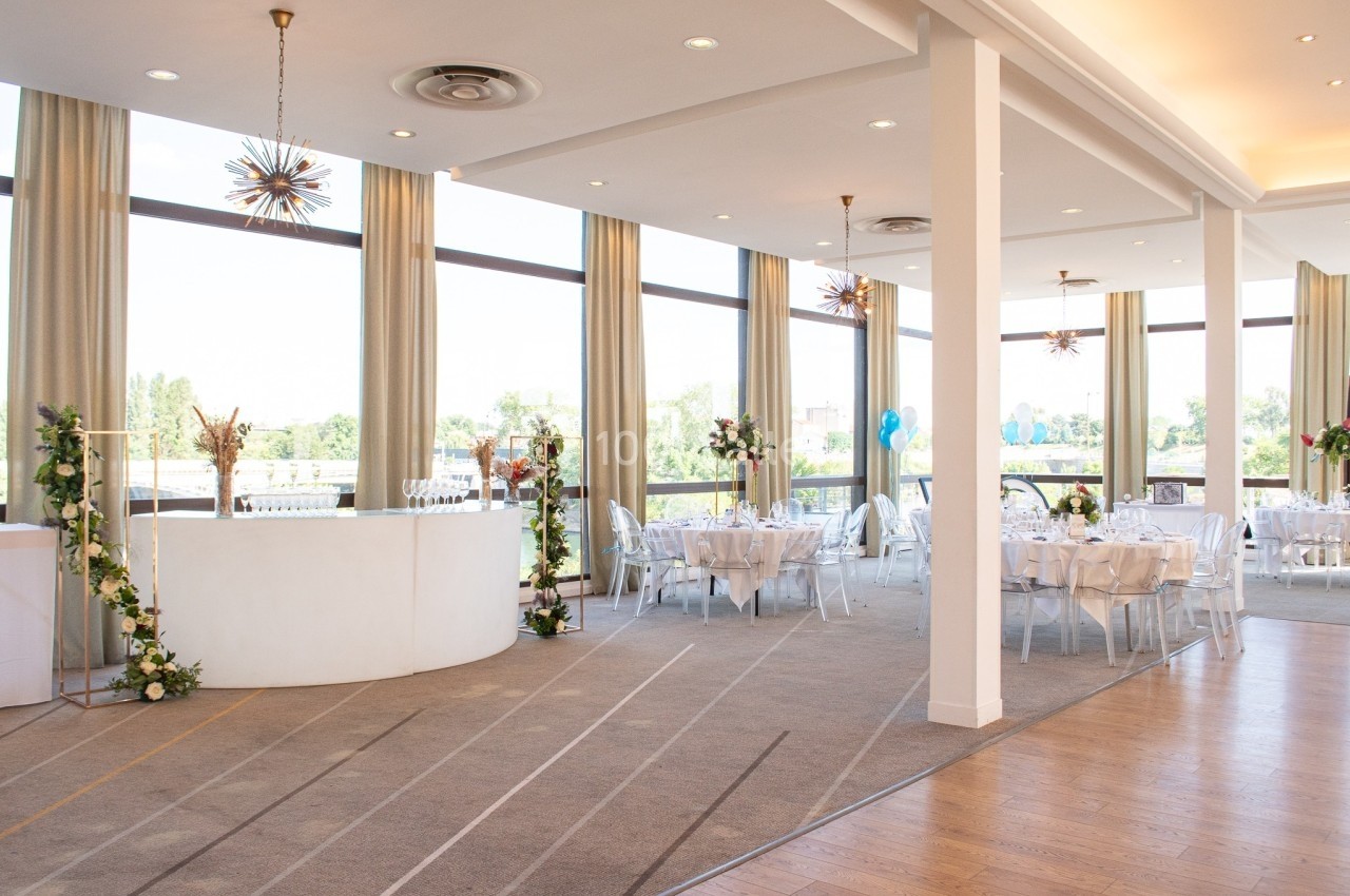 Salle lumineuse avec de grandes baies vitrées, tables dressées pour un événement et un bar décoré de fleurs.