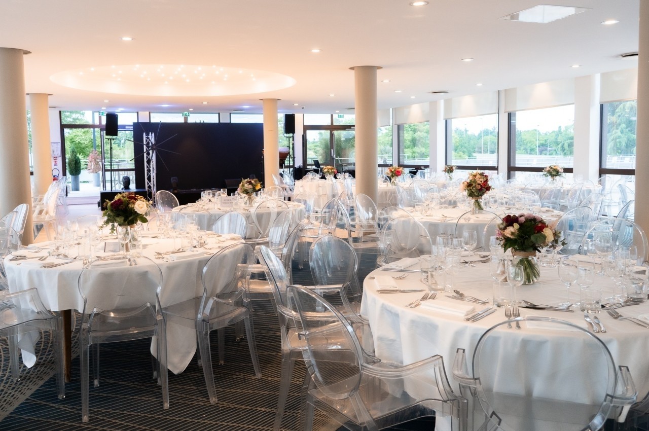 Salle de réception lumineuse avec tables rondes dressées, chaises transparentes et bouquets de fleurs au centre.
