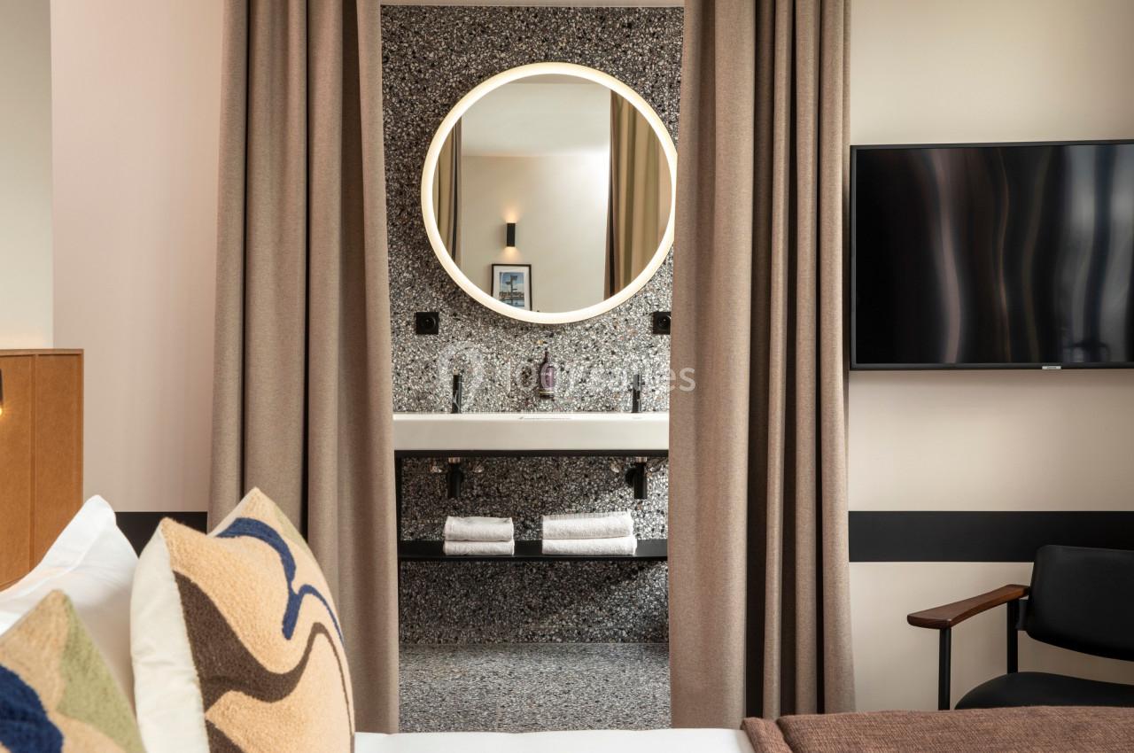 Vue d'une chambre d'hôtel avec un lit, un miroir rond éclairé et un lavabo en arrière-plan.