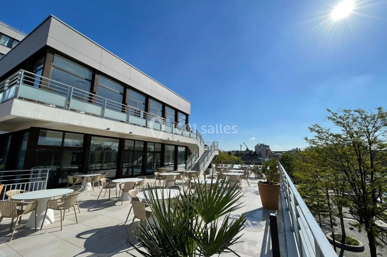 Terrasse ensoleillée avec tables et chaises, entourée de plantes, offrant une vue dégagée sur un ciel bleu.