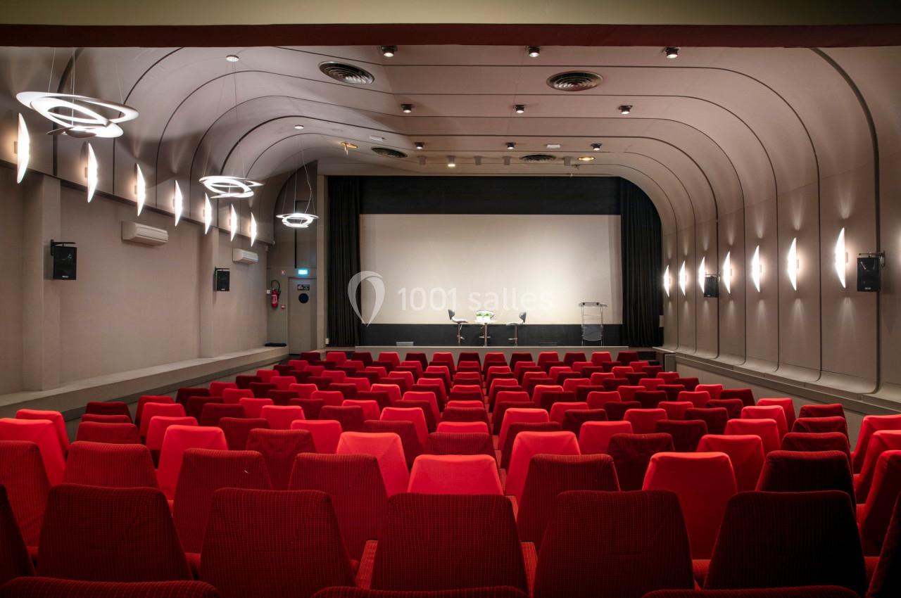 Salle de cinéma vide avec rangées de fauteuils rouges, écran blanc et éclairage moderne au plafond.