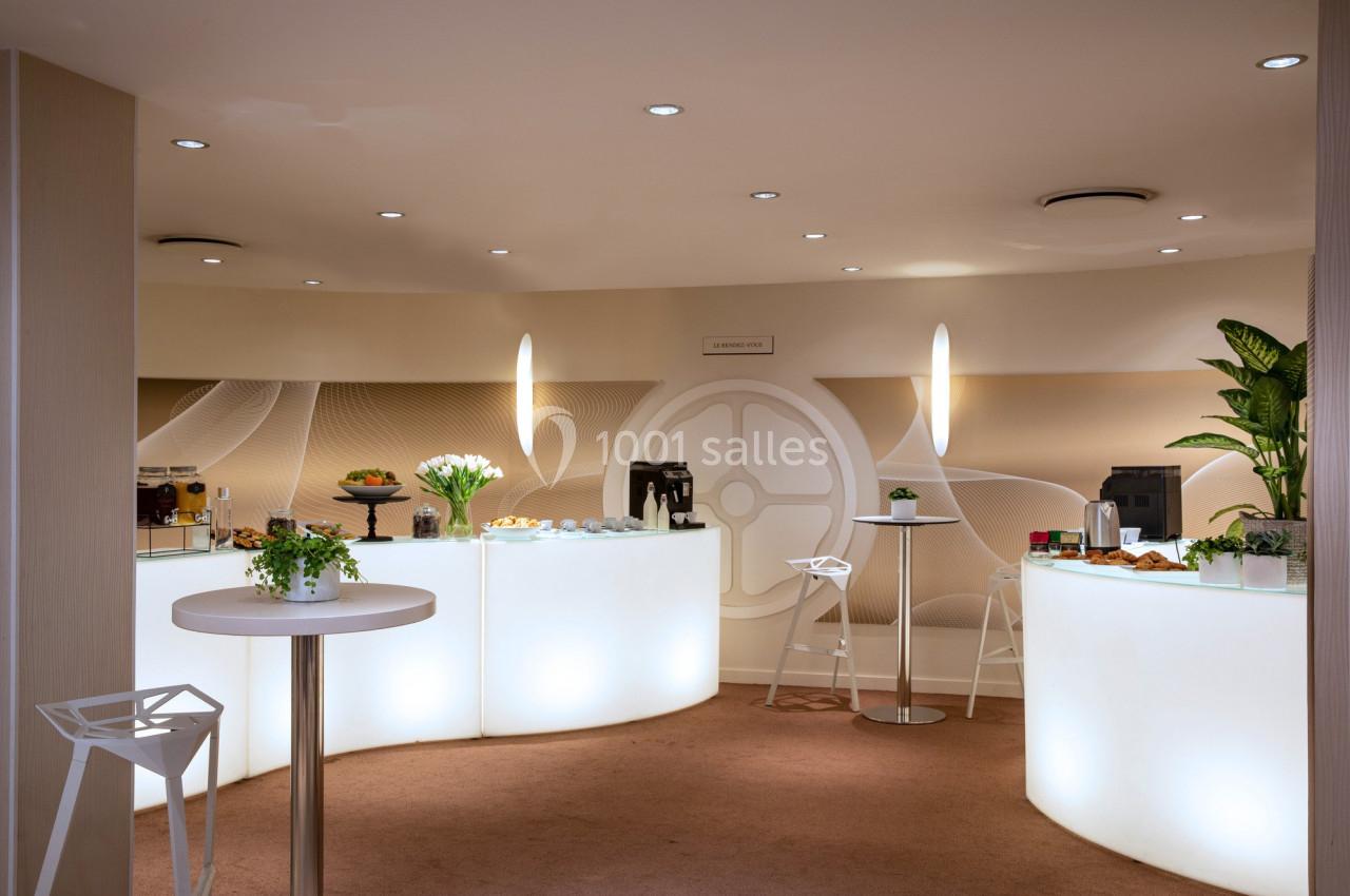 Espace lumineux avec tables hautes, plantes, machines à café et buffet disposés dans un cadre moderne et épuré.