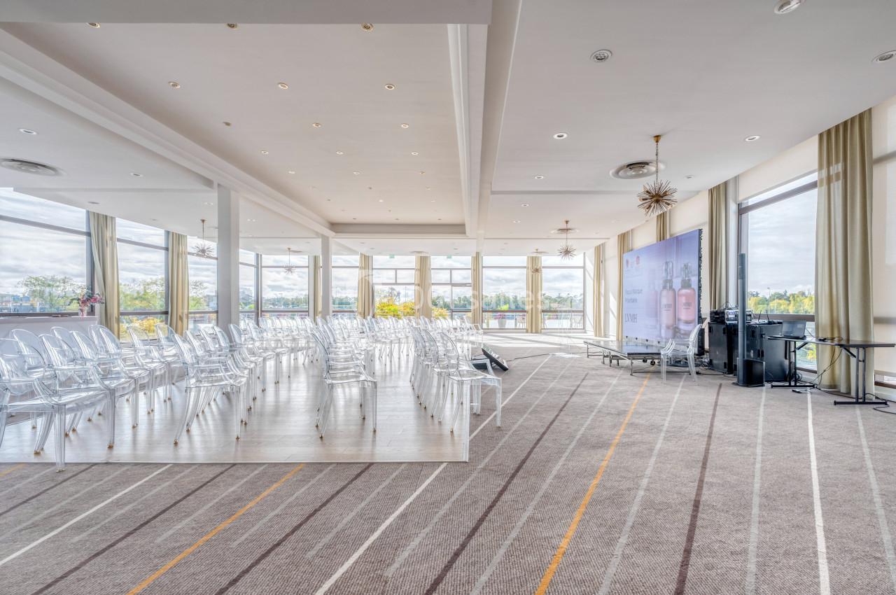 Salle lumineuse avec chaises transparentes alignées, grandes baies vitrées et équipement audio-visuel en fond.