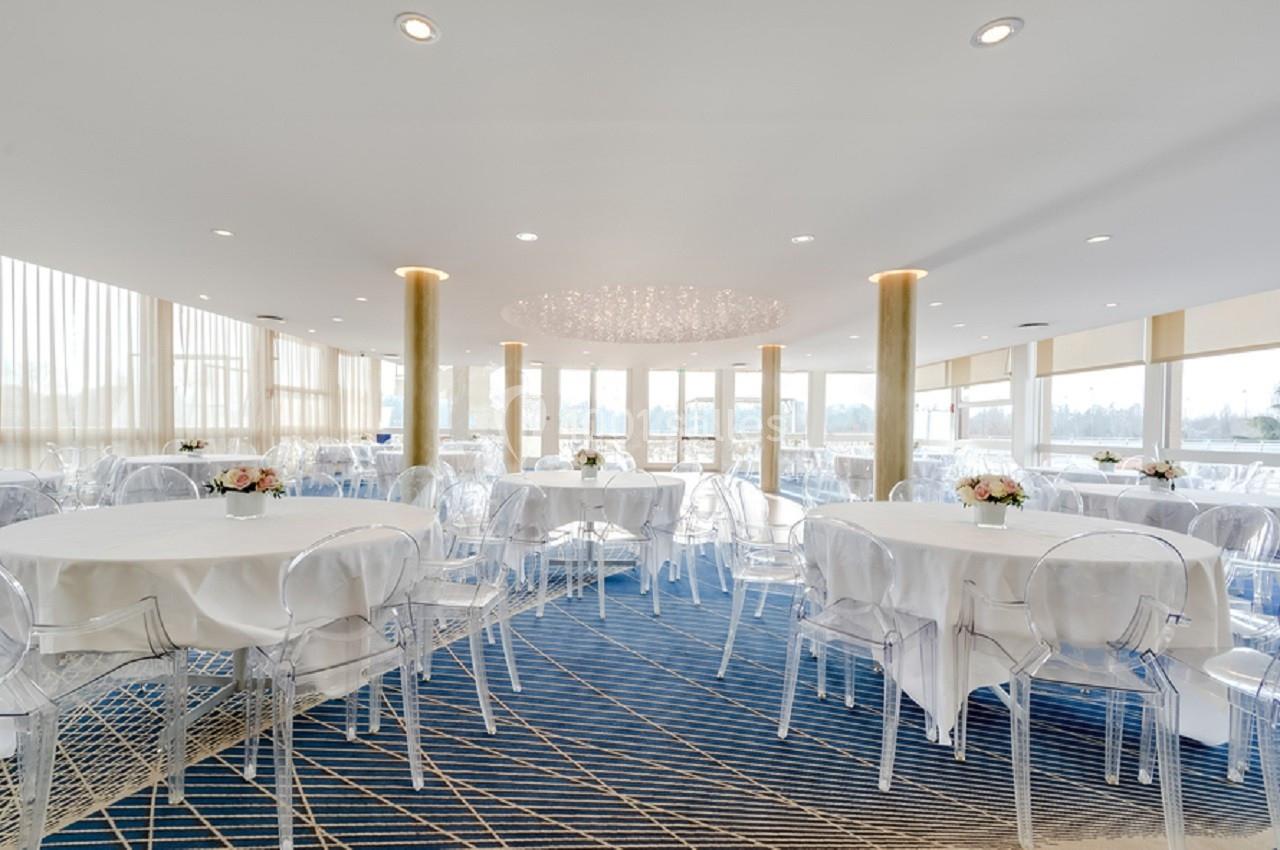 Salle lumineuse avec tables rondes dressées, chaises transparentes et grandes baies vitrées donnant sur l'extérieur.