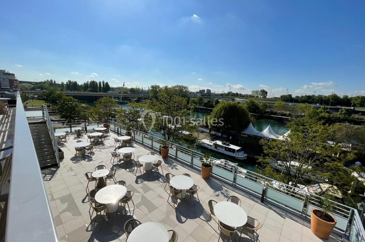Terrasse avec tables et chaises donnant sur une rivière bordée d'arbres, sous un ciel dégagé et ensoleillé.