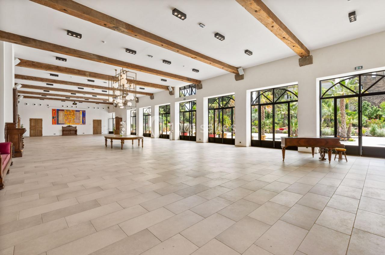 Salle spacieuse avec un sol en carrelage clair, poutres apparentes et grandes baies vitrées donnant sur un jardin.