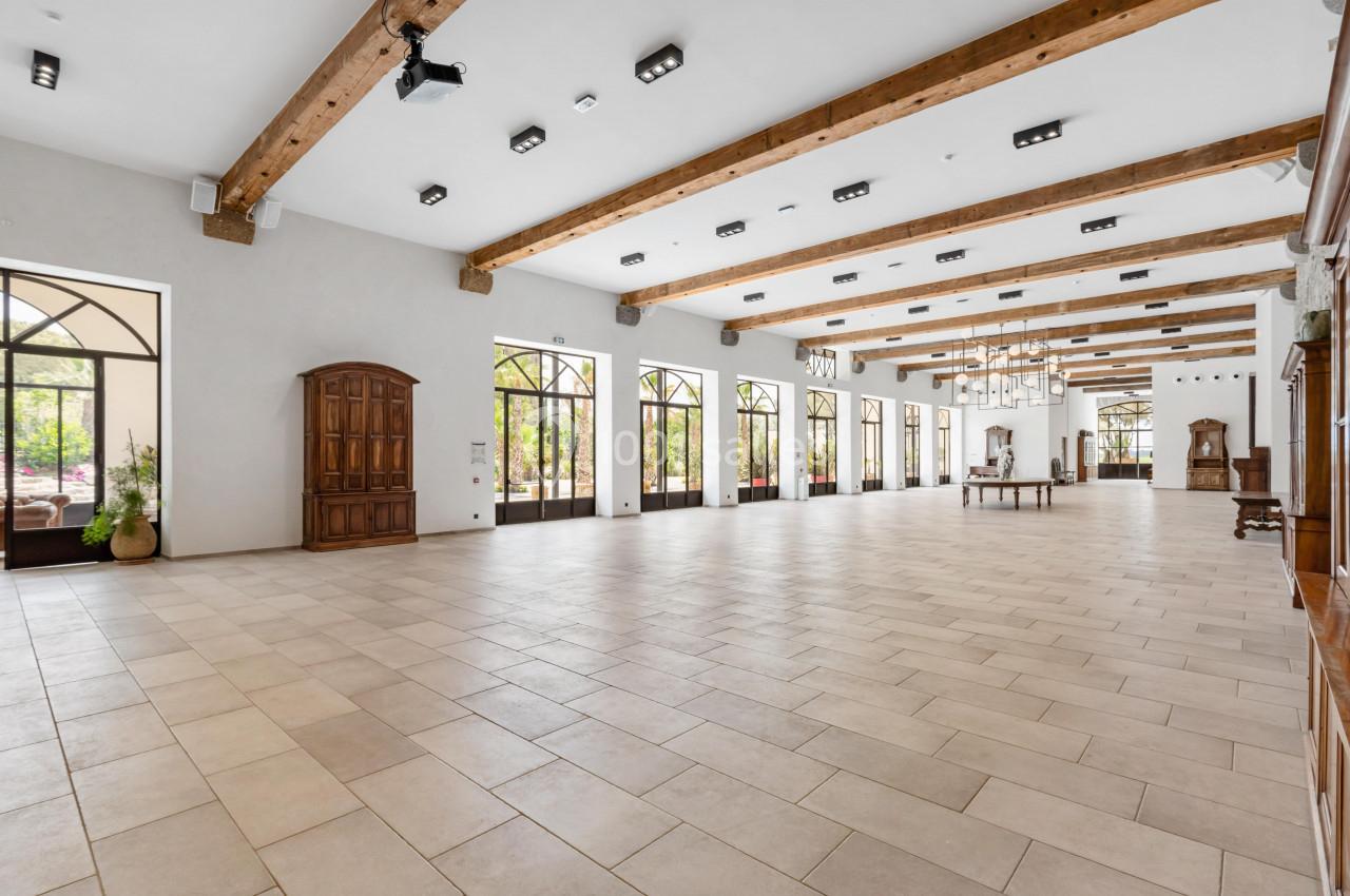 Grande salle lumineuse avec sol carrelé, poutres apparentes au plafond et grandes fenêtres donnant sur l'extérieur.