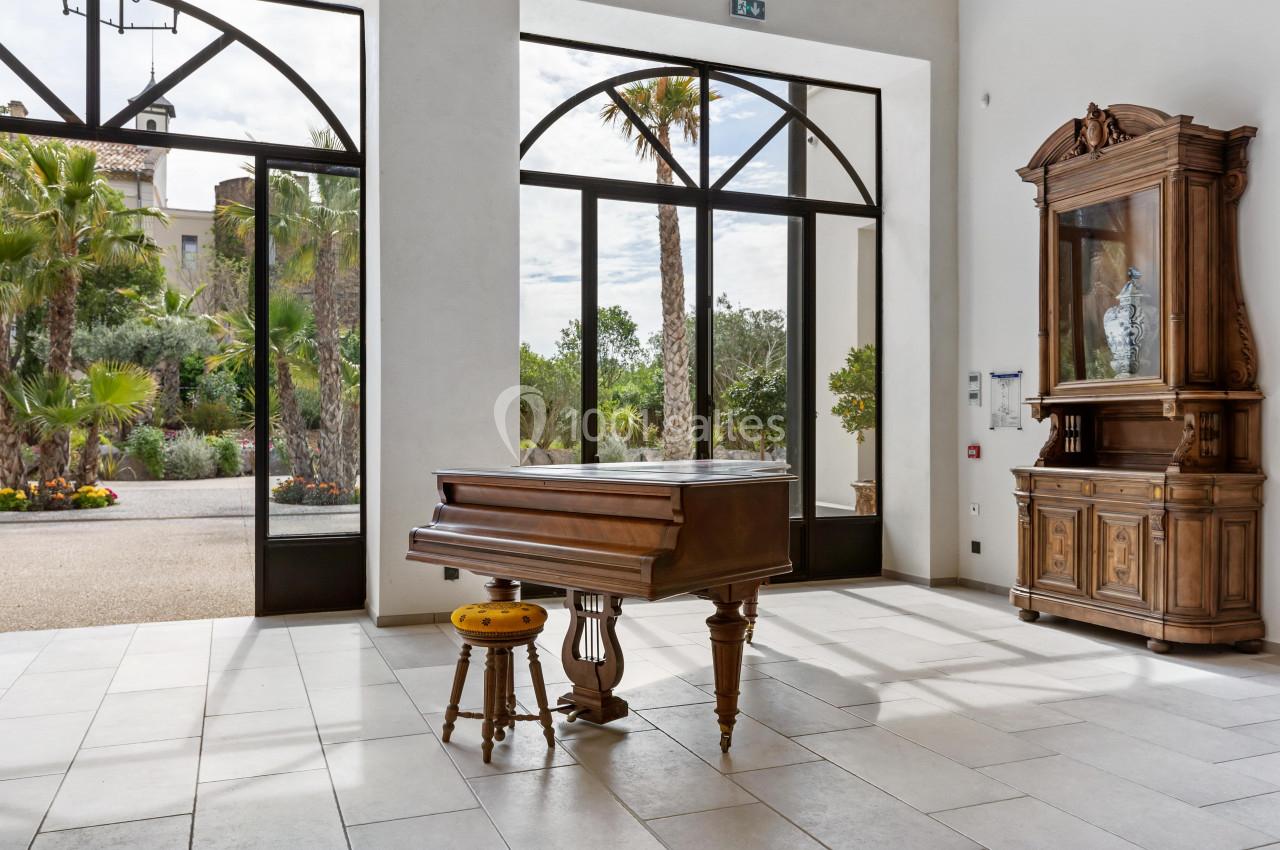 Piano ancien en bois avec tabouret, placé dans une pièce lumineuse aux grandes baies vitrées donnant sur un jardin.