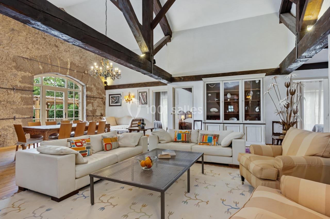 Salon spacieux avec poutres apparentes, canapés clairs, table basse, tapis décoratif et grande table à manger en arrière…