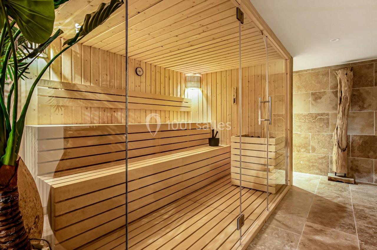 Sauna en bois clair avec bancs, seau et louche, éclairé par une lampe, entouré de murs en pierre beige.