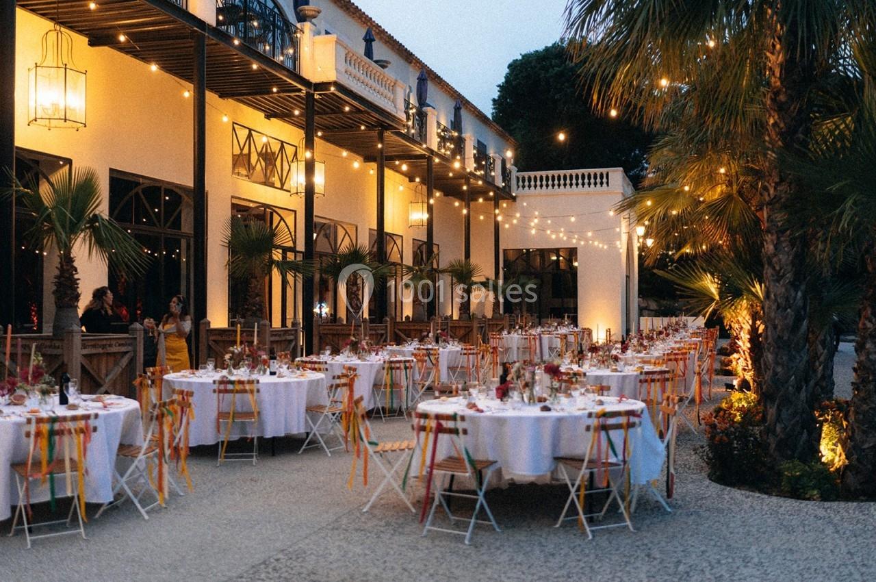 Tables décorées pour un dîner en extérieur, entourées de palmiers et éclairées par des guirlandes lumineuses au crépuscule.
