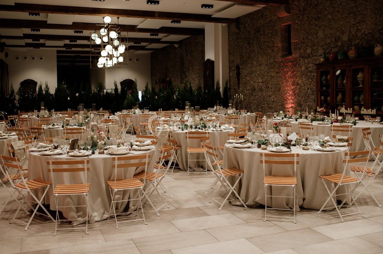Salle de réception décorée avec des tables rondes dressées, des chaises en bois et des éclairages suspendus.
