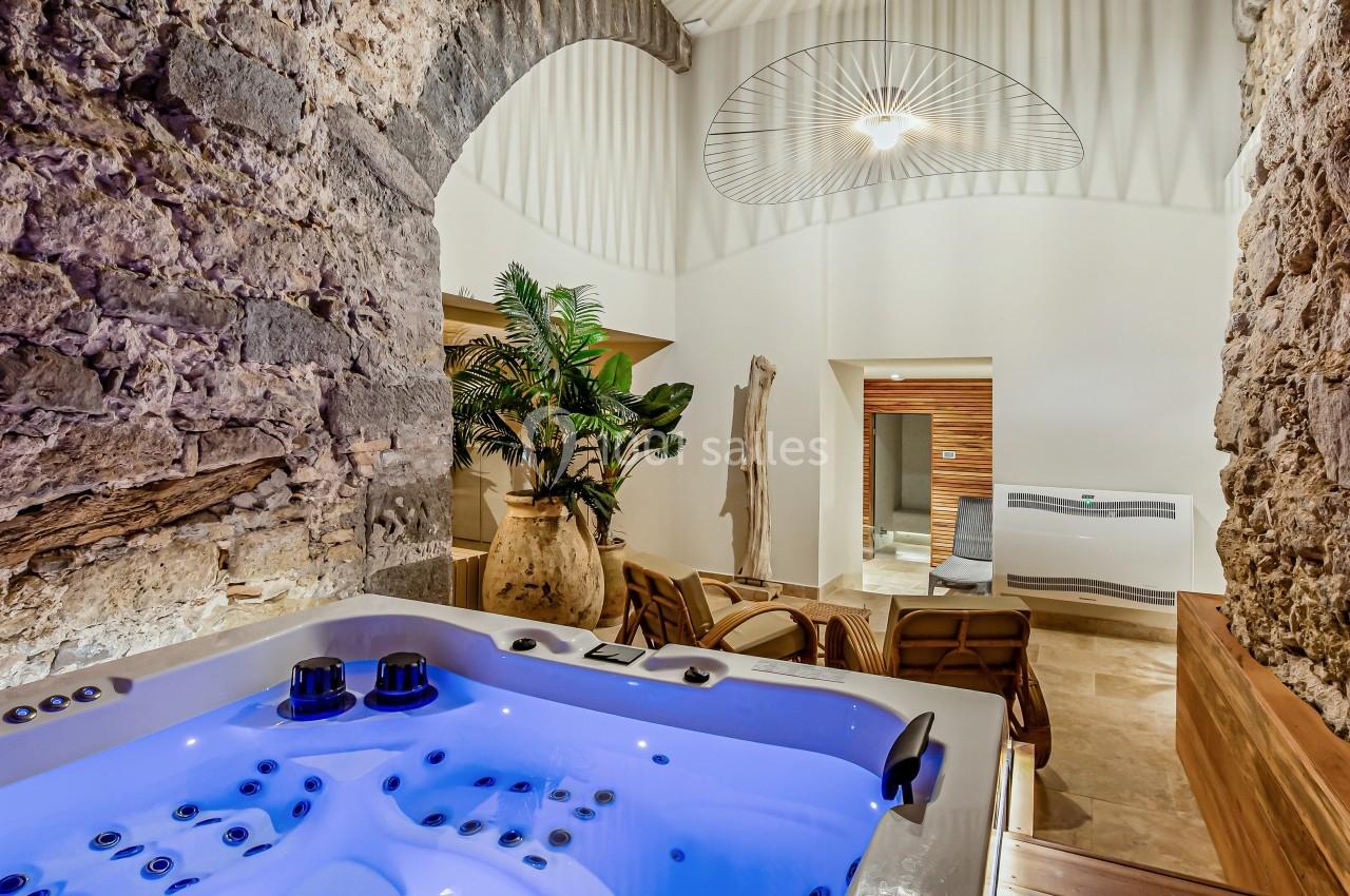 Jacuzzi éclairé dans une pièce voûtée avec murs en pierre, plantes décoratives et chaises longues en arrière-plan.