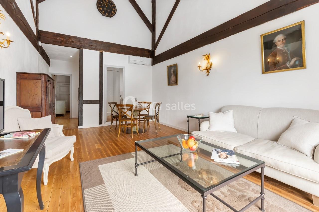 Salon lumineux avec poutres apparentes, canapé clair, table basse en verre, chaises en bois et décorations murales.