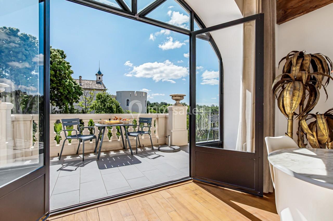 Vue depuis une pièce lumineuse sur une terrasse aménagée, avec table et chaises, donnant sur un paysage arboré et une tour.