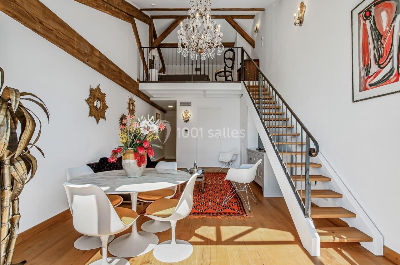 Salle à manger lumineuse avec table ronde, chaises blanches, poutres apparentes, escalier en bois et décoration murale.
