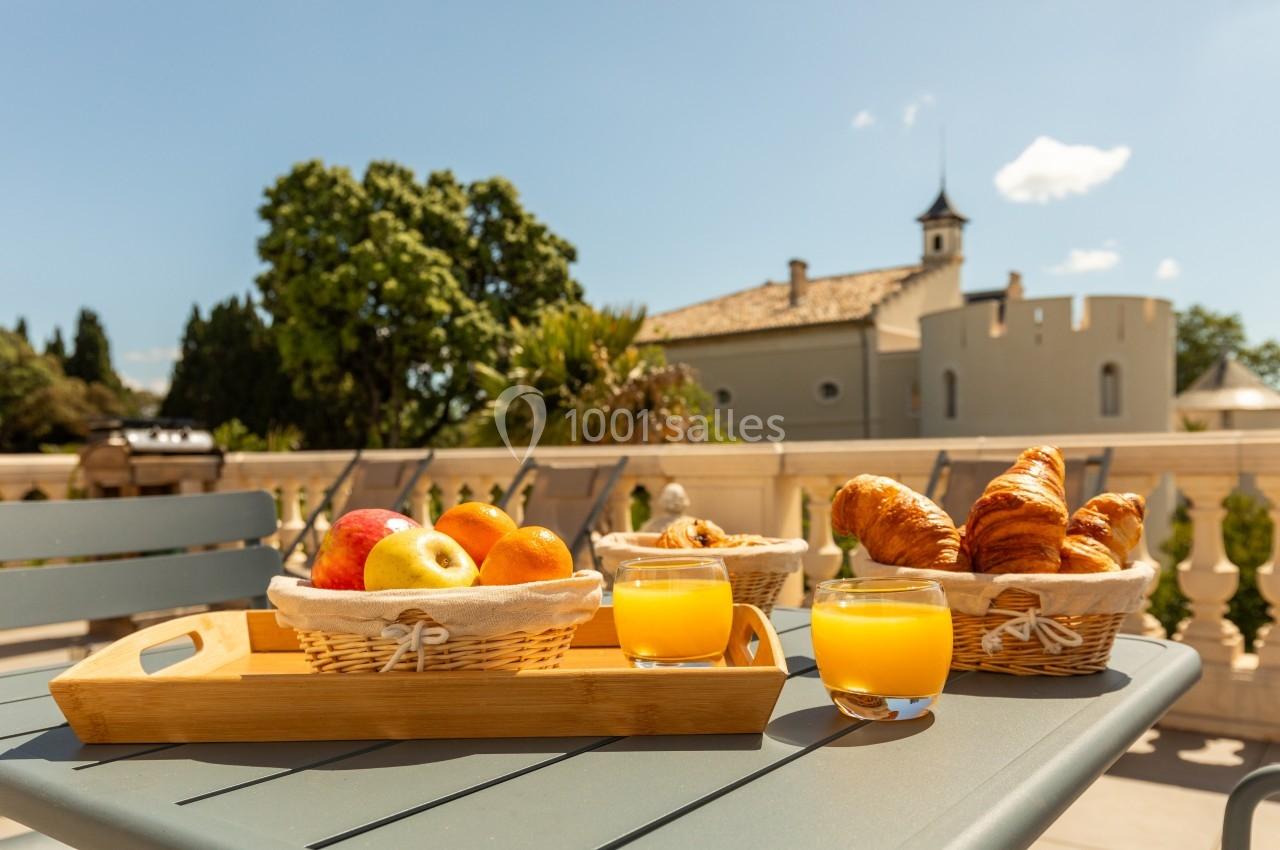Petit-déjeuner avec croissants, fruits et jus d'orange sur une table en terrasse, vue sur un bâtiment et des arbres.