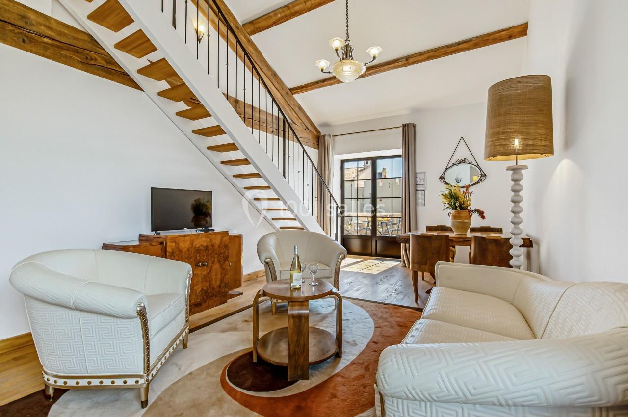 Salon lumineux avec mobilier classique, escalier en bois, plafond à poutres apparentes et grande fenêtre donnant sur l…