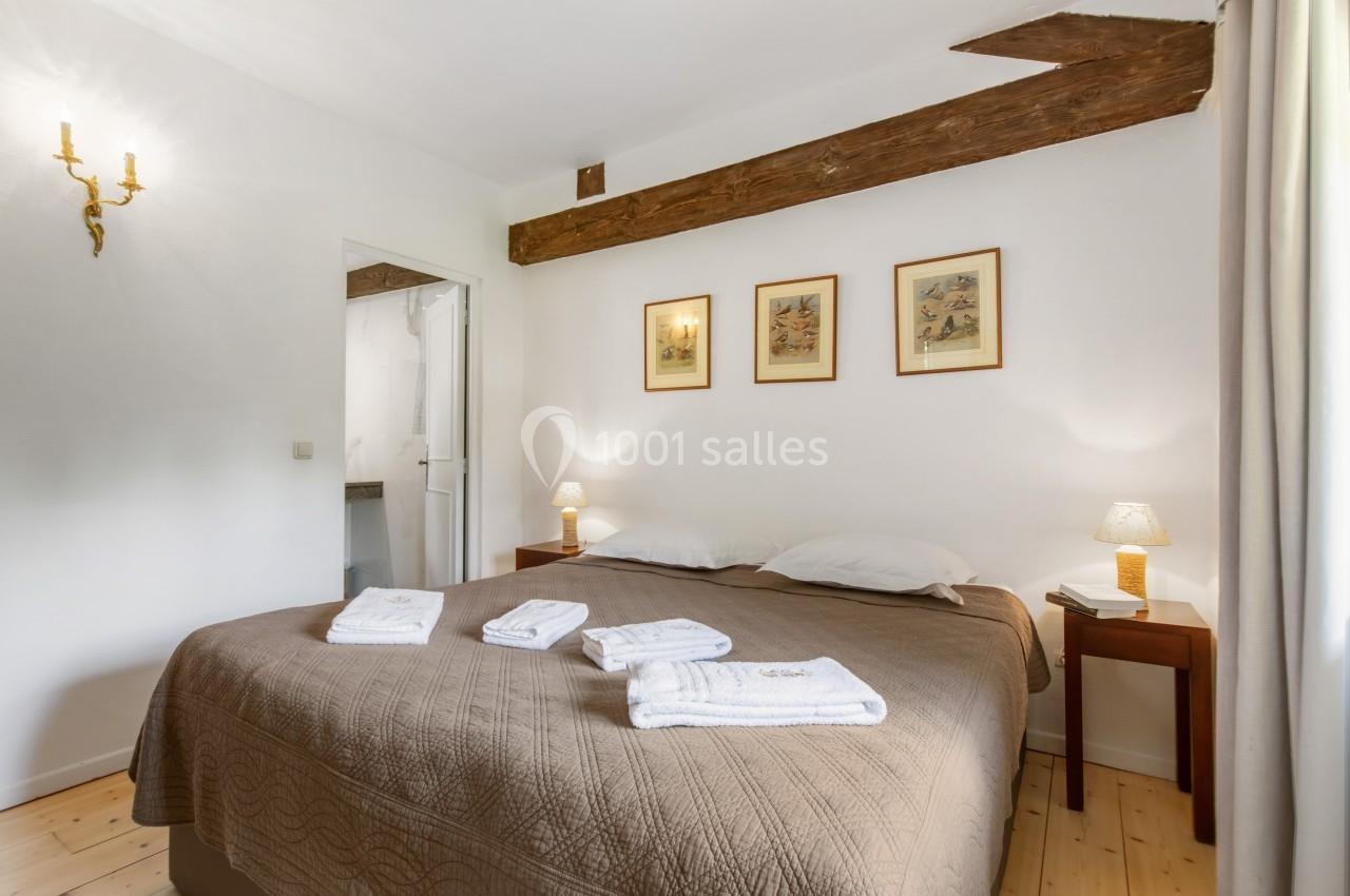 Chambre lumineuse avec lit double, poutres apparentes, décor sobre et linge de lit plié sur la couverture.