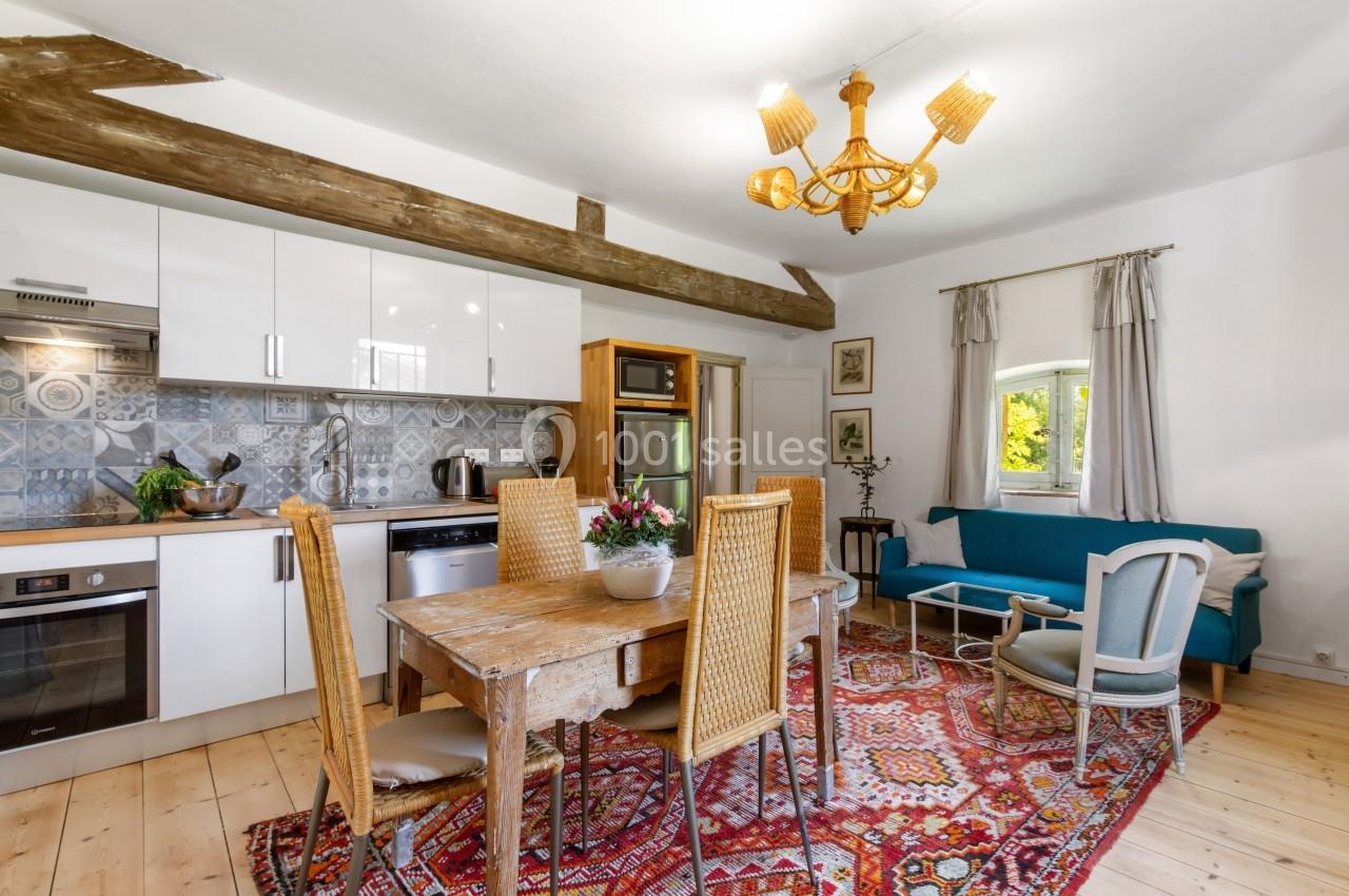 Cuisine ouverte avec table en bois, chaises en rotin, tapis coloré, canapé bleu et poutres apparentes.