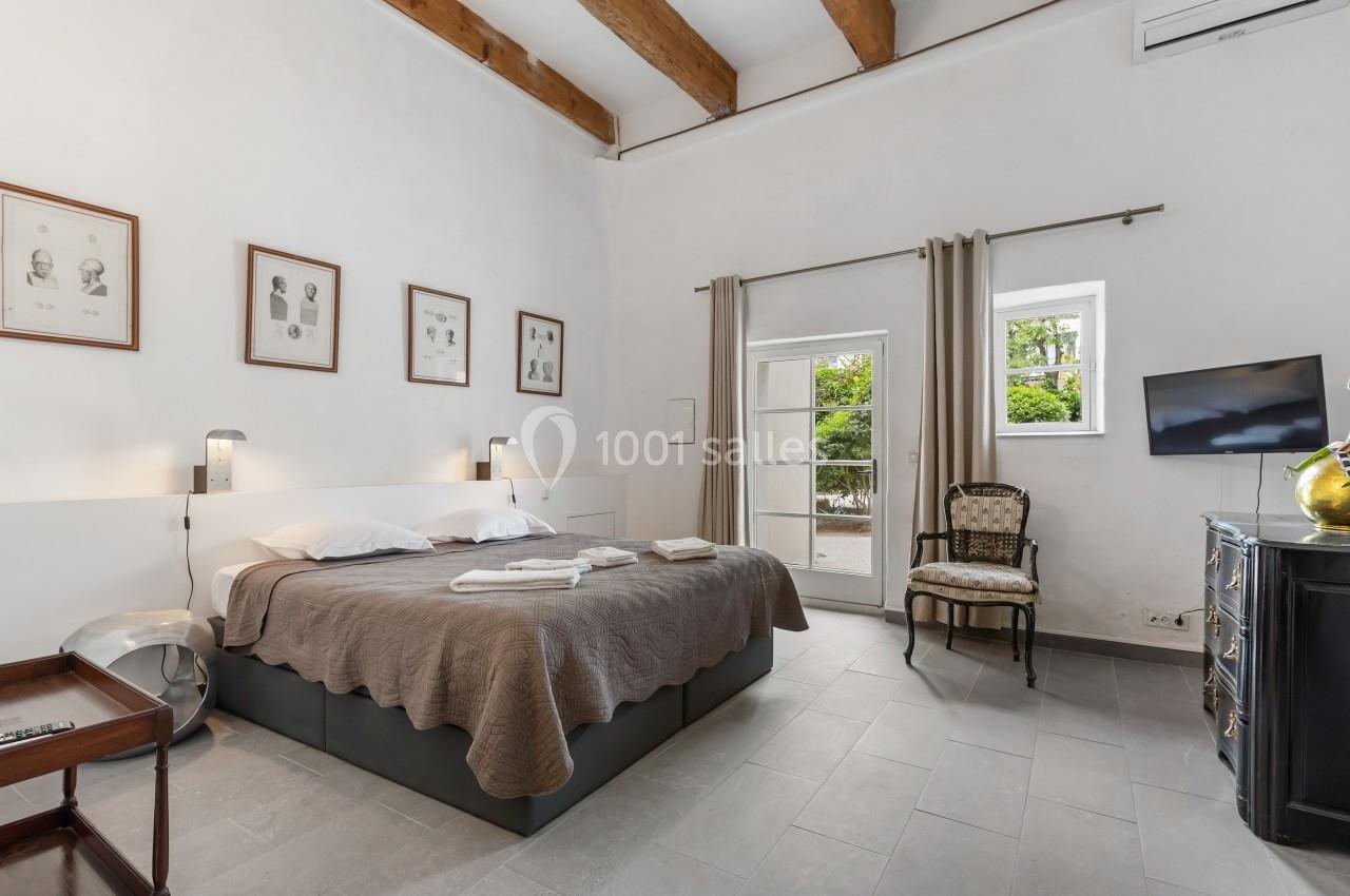 Chambre lumineuse avec lit double, murs blancs, poutres apparentes, fenêtres donnant sur un jardin et mobilier classique.