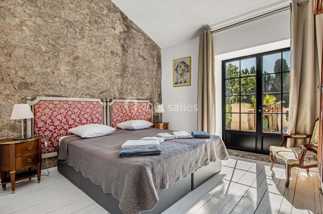 Chambre lumineuse avec un lit double, murs en pierre, grandes fenêtres donnant sur un balcon et mobilier classique.