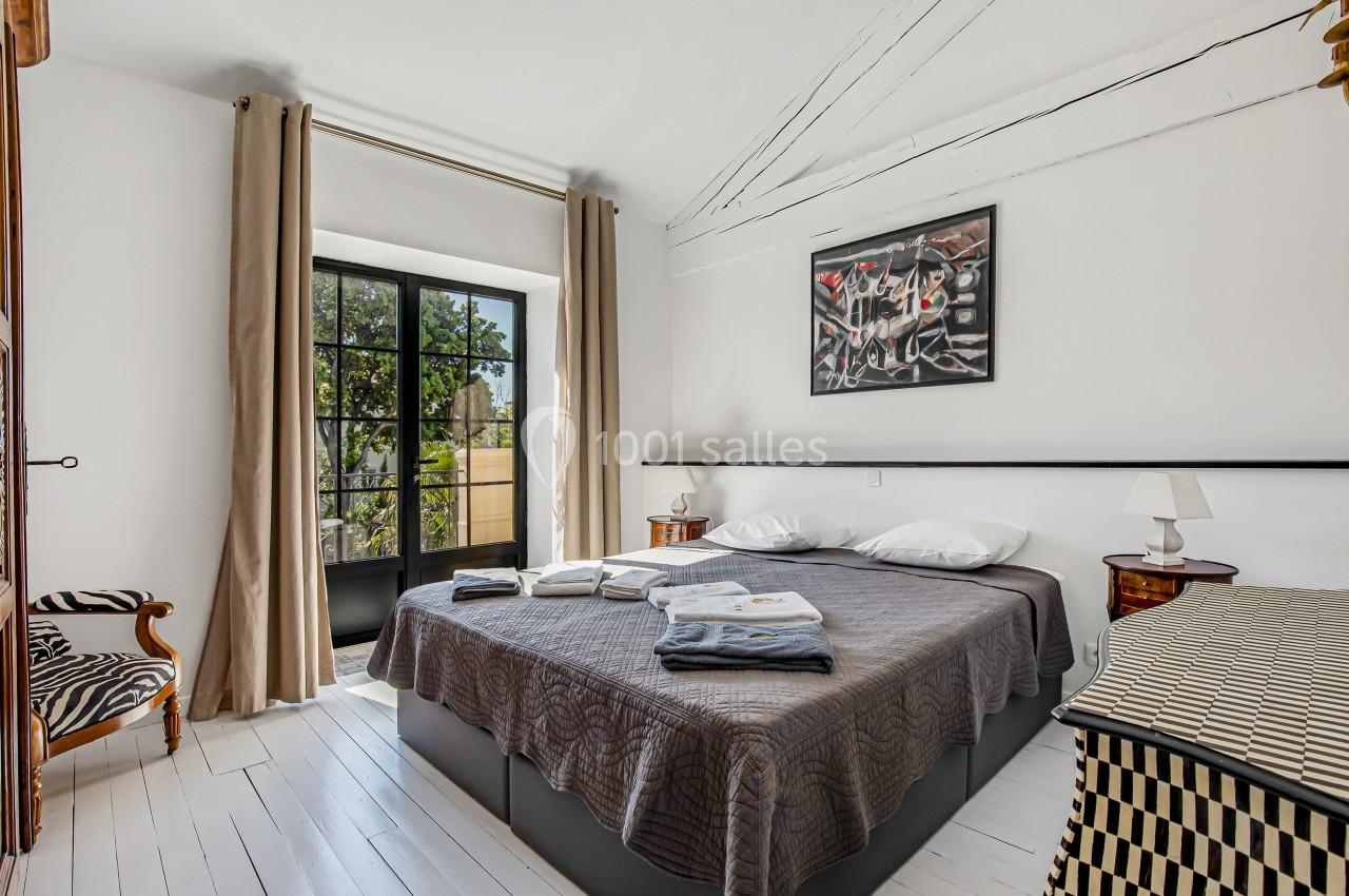 Chambre lumineuse avec lit double, draps pliés, fauteuil rayé, grande fenêtre donnant sur un jardin.