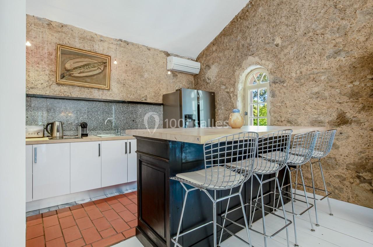 Cuisine avec murs en pierre, comptoir central noir, tabourets métalliques et fenêtre en arche laissant entrer la lumière.