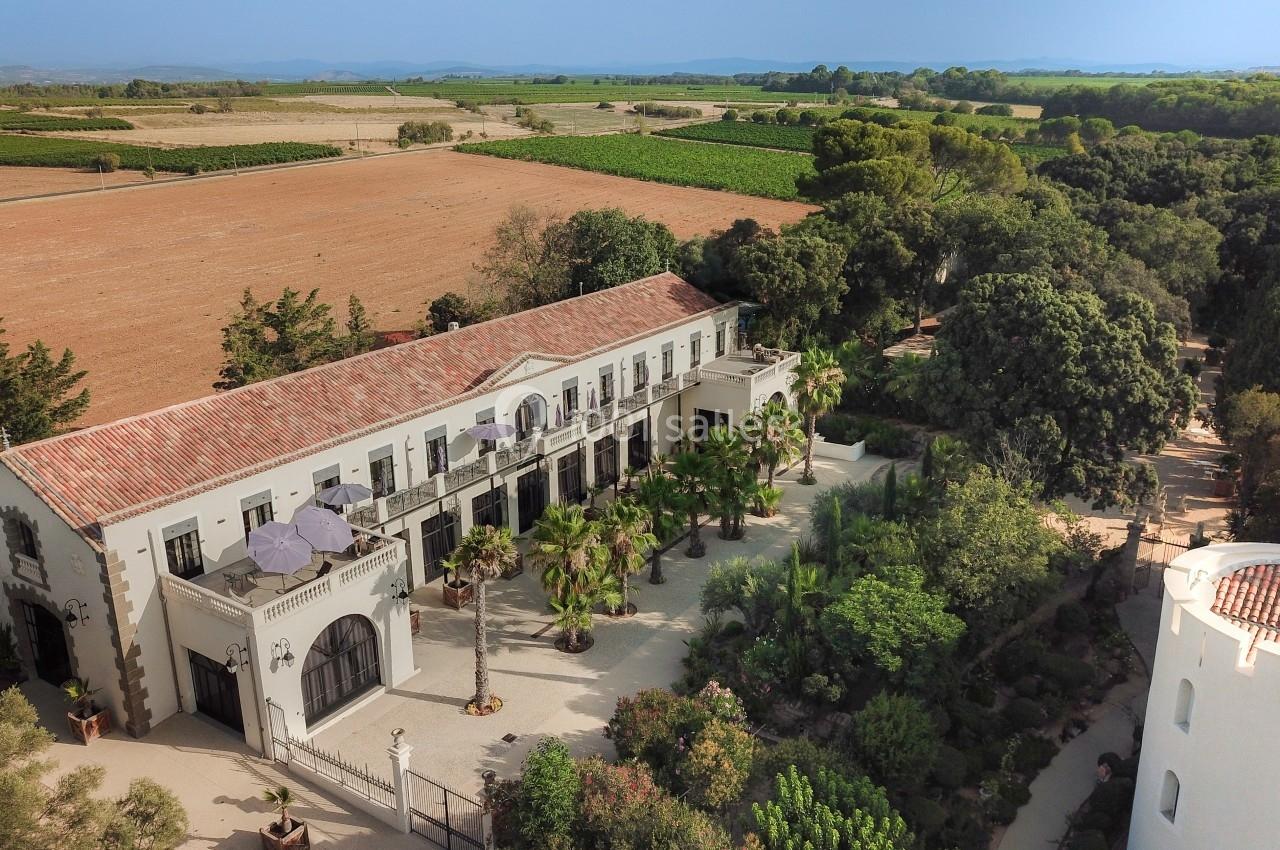 Vue aérienne d'un bâtiment méditerranéen entouré de jardins, avec des champs et des collines à l'arrière-plan.