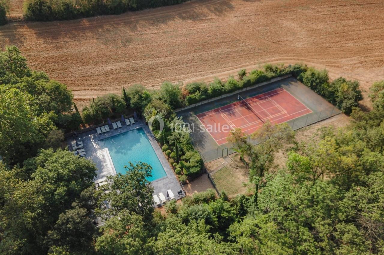 Vue aérienne d'une piscine entourée de chaises longues et d'un terrain de tennis au milieu d'un paysage rural.
