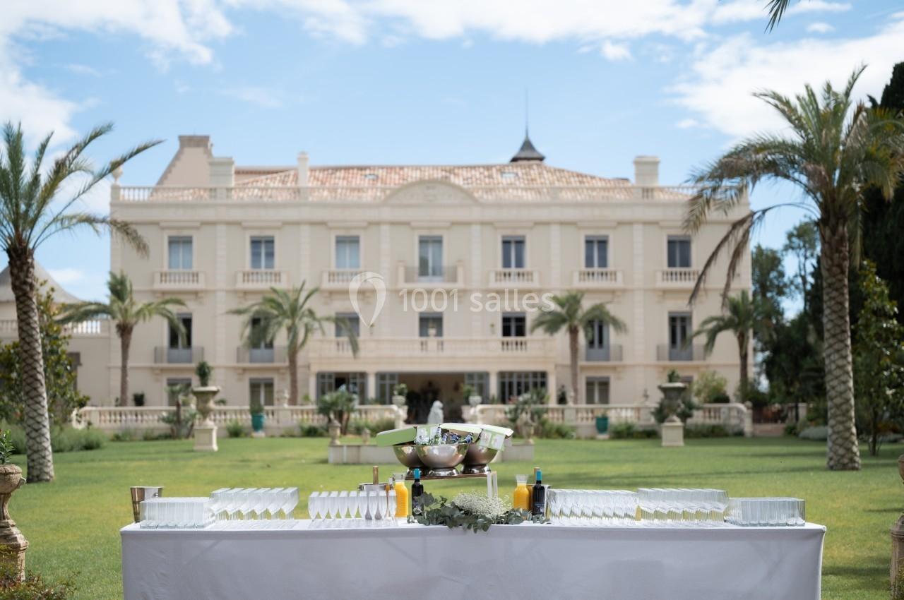 Table dressée avec verres et boissons devant un grand bâtiment élégant entouré de palmiers et de verdure.