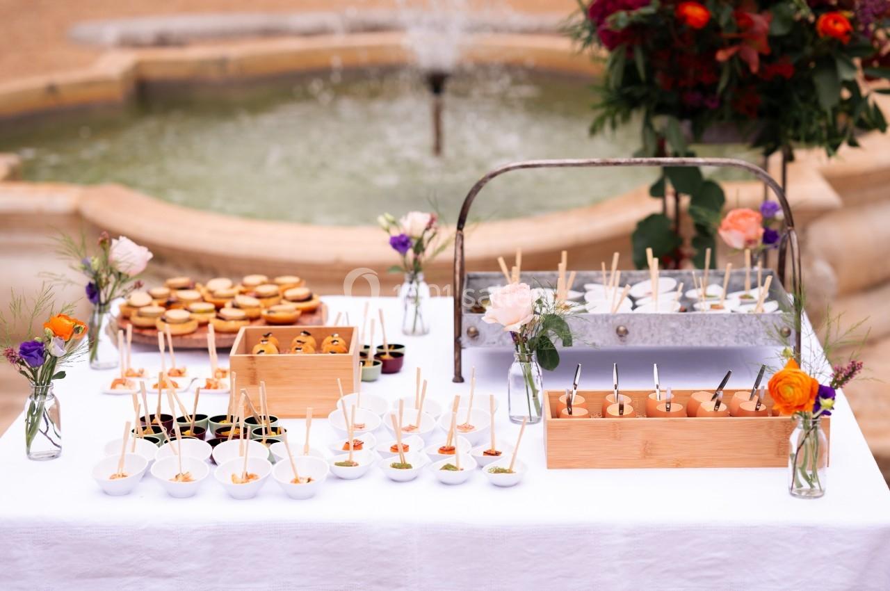Table dressée avec des amuse-bouches variés, présentés dans des contenants élégants, devant une fontaine en arrière-plan.