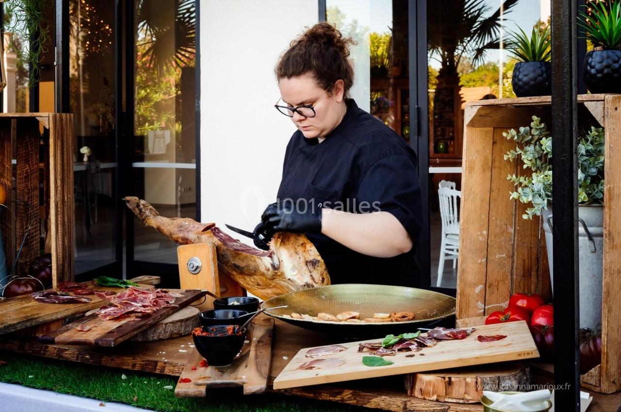 Une personne découpe un jambon sur un support en bois, entourée de planches garnies de charcuterie et de légumes.