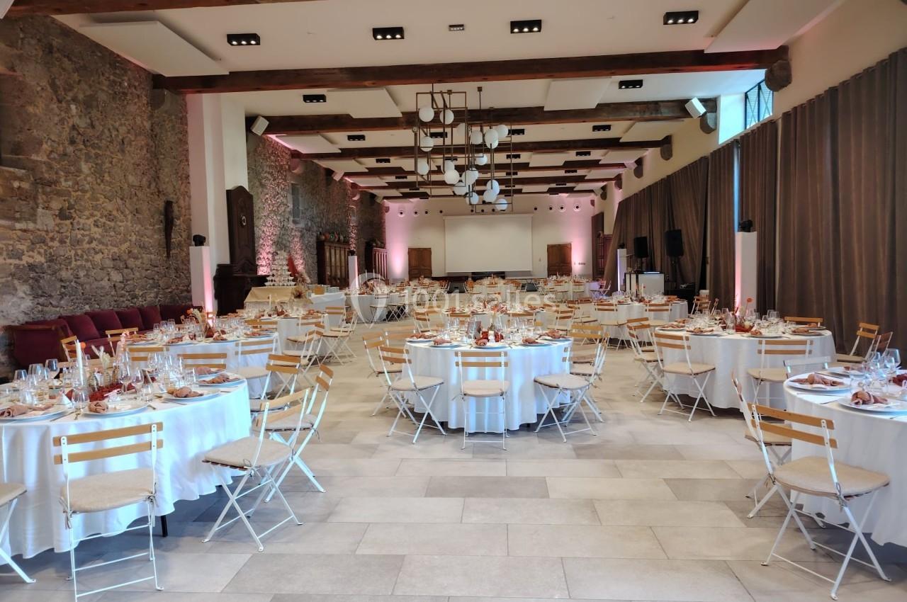 Salle de réception décorée avec des tables rondes dressées, chaises en bois et éclairage chaleureux.