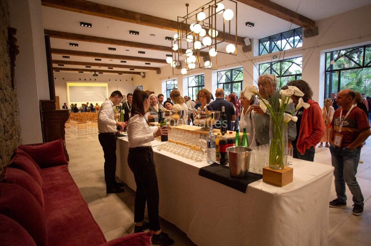 Des personnes se servent à un buffet avec boissons et verres, dans une salle lumineuse avec des tables et des lustres…