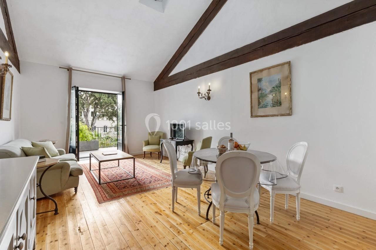 Salon lumineux avec parquet, poutres apparentes, canapé, table ronde, et grande baie vitrée donnant sur un balcon.