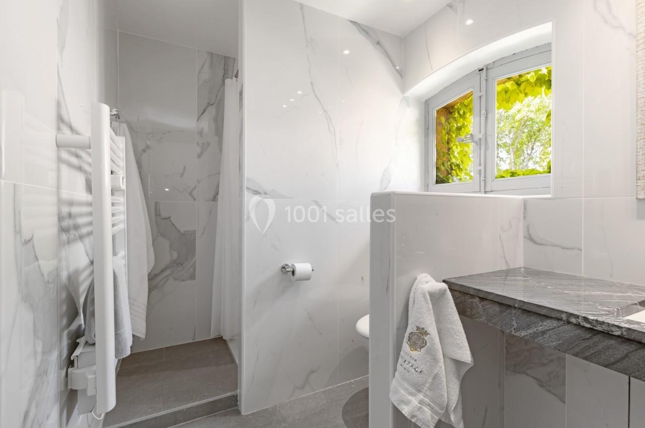 Salle de bain moderne avec murs en marbre, douche ouverte, lavabo en pierre et fenêtre donnant sur des feuillages.