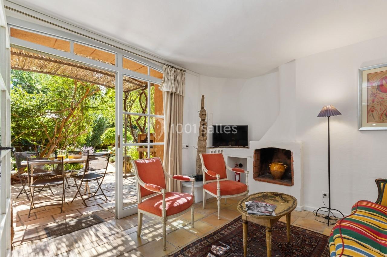 Salon lumineux avec cheminée, deux fauteuils, table basse et vue sur une terrasse ombragée avec table et chaises.