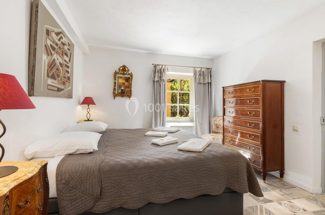 Chambre lumineuse avec lit double, commode en bois, fauteuil et fenêtre donnant sur un jardin.